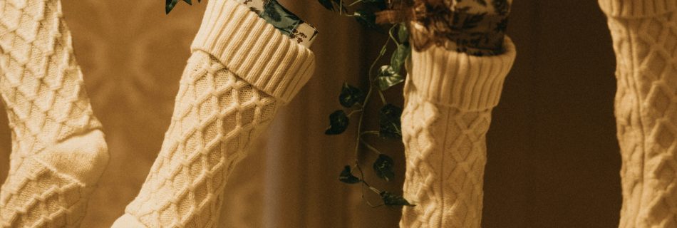Four cream, cable-knit Christmas stockings hung on a garland, partially filled with small gifts.