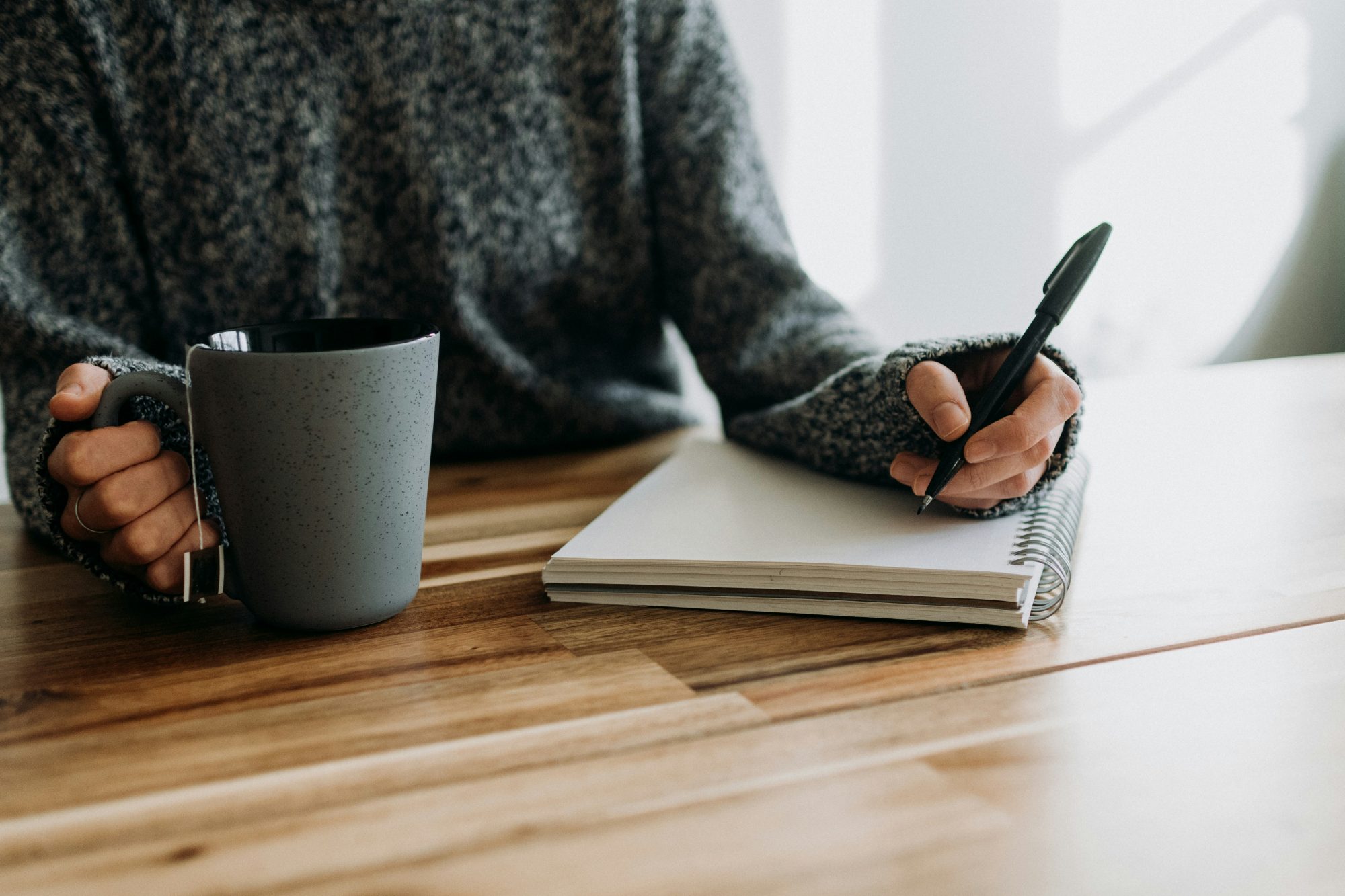 A person in a grey sweater sitting at a wooden table, holding a mug of tea and writing in a spiral notebook with a black pen.
