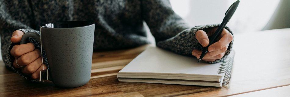 A person in a grey sweater sitting at a wooden table, holding a mug of tea and writing in a spiral notebook with a black pen.